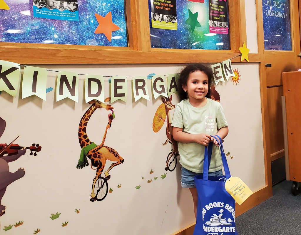 Bianca leans against the wall, holdering her 1000 Books Before Kindergarten bag. She has a shy smile, but is clearly proud of her accomplishment. She is about 3 years old, with light brown skin and curly hair. she is wearing a green shirt and jean shorts.