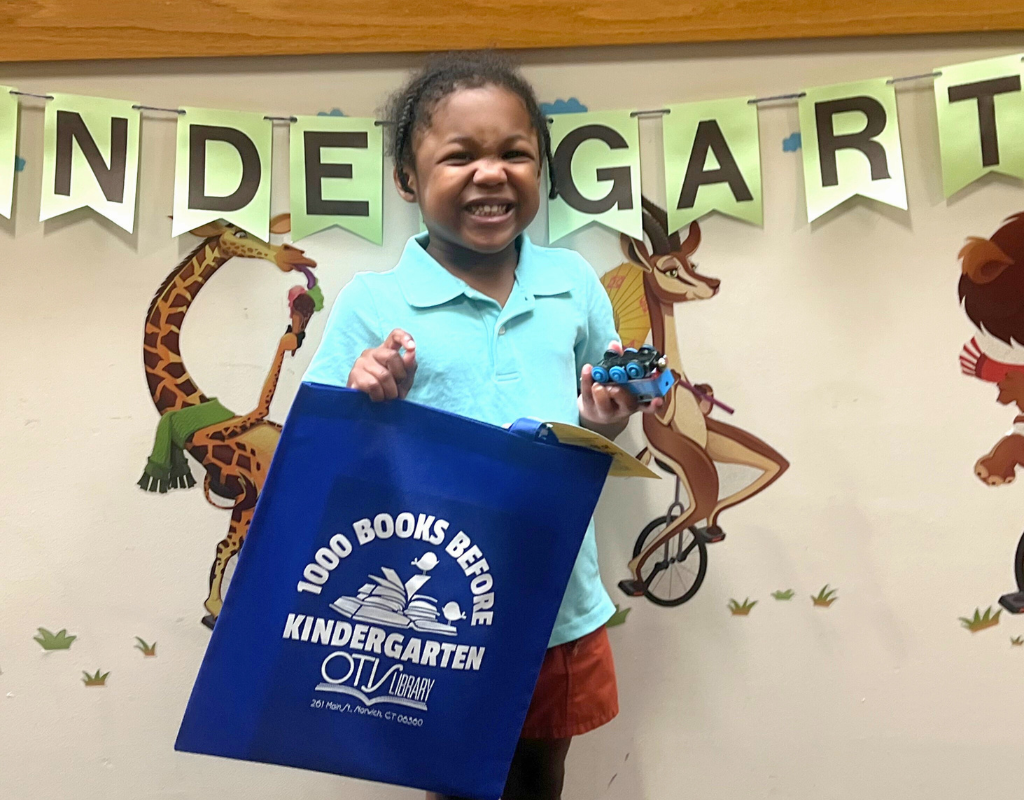 Everette's smile is beaming, stretching across his face, as he holds up a a bag labeled as having read 1000 Books Before Kindergarten. Everett is around 4 years old, black skin, with a blue shirt and red shorts. His joy and excitement is infectious.