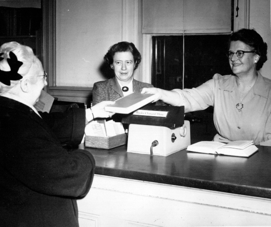 Librarians check out a book to a woman. The photo is undated, but the black and white image features fashions and styles popular in the 1950s.