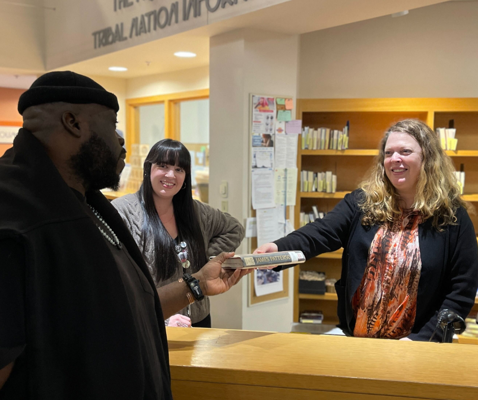 Two staff members are behind the desk. one hands a book to a man on the other side. They are all smiling at each other.