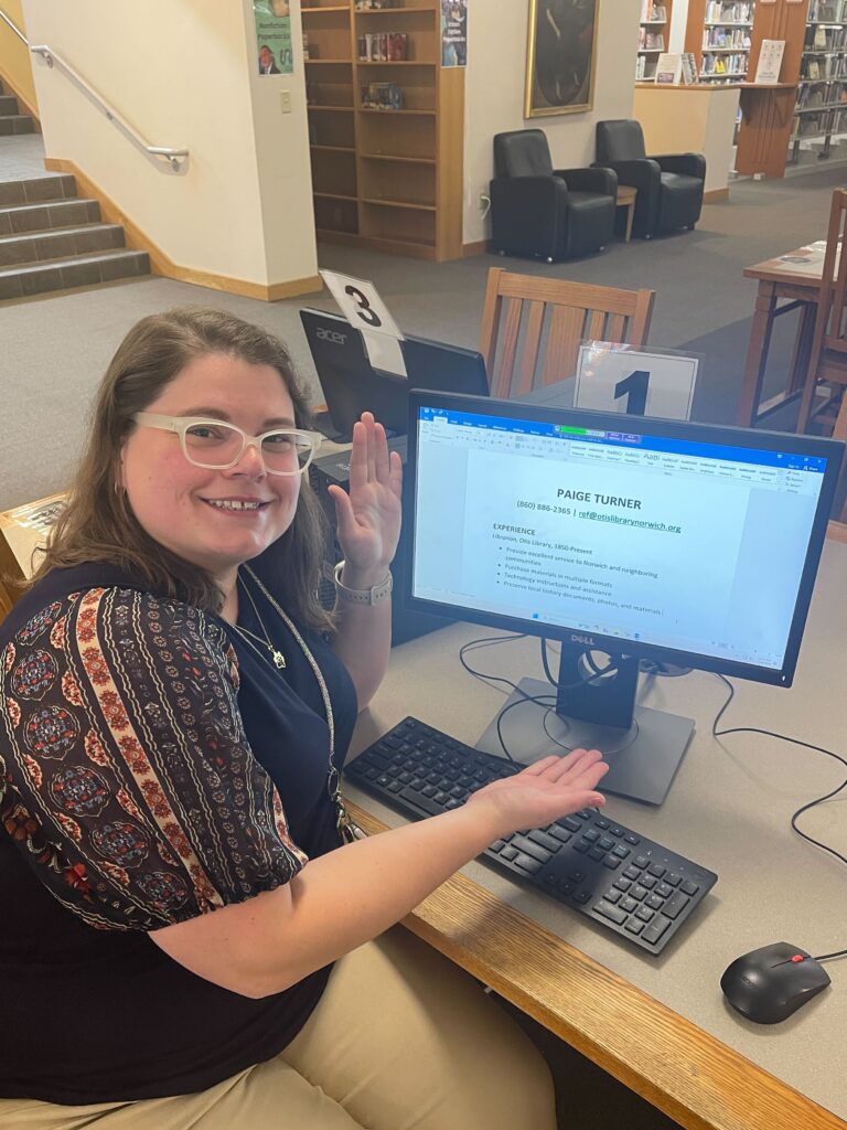 Jessa sits at a computer and gestures to the screen with a smile. The screen shows a resume template for a person named Paige Turner. Jessa likes puns.