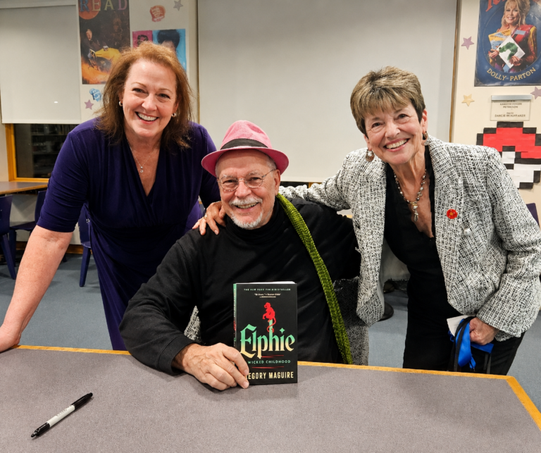 Three people smile together at a book signing event as author Gregory Maguire holds a copy of his book Elphie: A Wicked Childhood.
